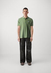 Camisa polo verde con tapeta de tres botones y pequeño logo en el pecho, combinada con pantalones negros de corte holgado. El modelo está de pie frente a un fondo liso.
