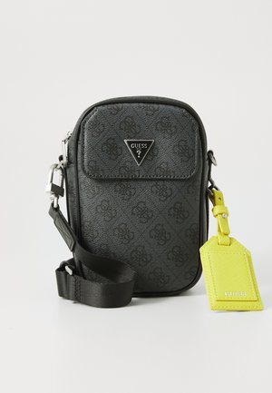 Schwarze Guess Umhängetasche mit gemustertem Textil, Vorderklapptasche, verstellbarem Riemen und angehängtem leuchtend gelben Guess Gepäckanhänger.