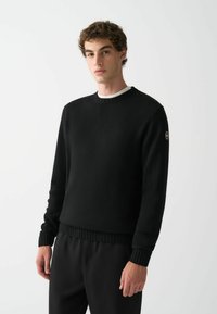 Schwarzer Rundhals-Pullover aus Strickstoff mit gerippten Bündchen und Saum. Mit einem kleinen Logopatch am linken Ärmel.