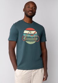 Türkises Baumwoll-T-Shirt mit Grafiken eines Retro-Fahrrads in Rot, Orange und Mintgrün auf einem kreisförmigen Hintergrund mit horizontalen Streifen.