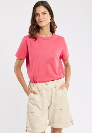 Femme portant un T-shirt corail à manches courtes, rentré dans un short beige taille haute avec de grandes poches devant, debout les mains dans les poches.