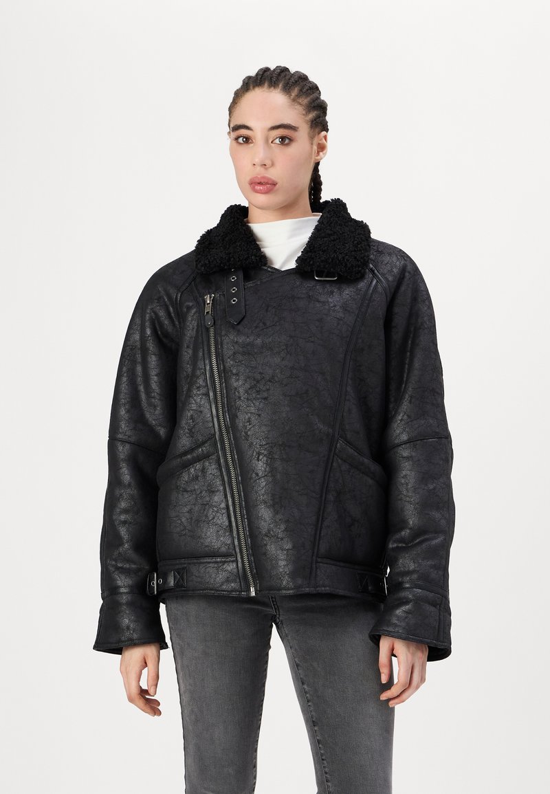 Juodas dirbtinės odos švarkas su pūkuotu apykakle, užtrauktuku ir šoninėmis kišenėmis. Textūruotas paviršius ir laisvas, per didelis fasonas.