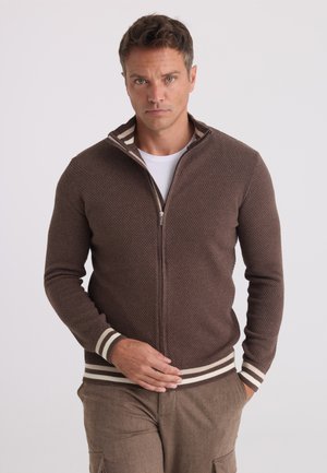 Homme portant un pull à fermeture éclair marron texturé avec des poignets et un ourlet rayés, et un pantalon marron, debout avec une main sur la fermeture éclair.