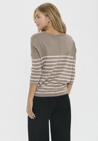 Una donna bionda di spalle indossa un maglione taupe con strisce bianche orizzontali sulle maniche e sul retro, abbinato a pantaloni neri larghi.