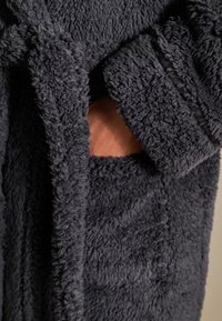 Antracietgrijze, pluizige badjas met een zachte textuur. Voorzien van een ceintuurlint en een zijzak waarin een hand steekt.