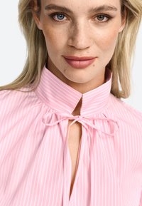 Blusa rosa con colletto e strisce bianche verticali, presenta un dettaglio con fiocco al collo e tessuto arricciato attorno al colletto. Tessuto morbido.