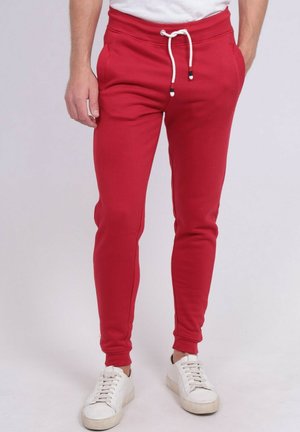 Homme portant un pantalon de jogging rouge ajusté avec un cordon blanc et des baskets blanches, debout devant un fond clair uni.