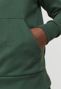 Sudadera de color verde oscuro con bolsillo canguro, puños y dobladillo acanalados. Textura de tela suave, con un corte clásico y un diseño minimalista.