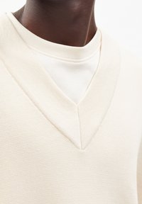 Maglione color crema a v con tessuto a costine, indossato sopra una camicia beige chiaro. Il collo presenta un taglio pulito e angolare.