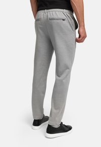 Mies, joka käyttää vaaleanharmaita rentoja housuja takana vetoketjutaskuilla ja mustia lenkkareita, seisoo yhdellä kädellä sivutaskun lähellä.