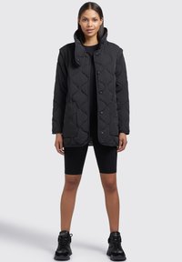Veste matelassée noire avec un col montant, des boutons-pression et des poches latérales, présentant une texture lisse et un design mi-long.