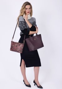 To håndtasker vises: en mørkerød læder tote med et cirkulært logo og en mønstrede burgundy og guld clutch. Modellen har en sort strikket kjole på.