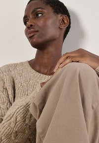 Beiger Strickpullover mit einem Zopfmuster, kombiniert mit leicht braunem Stoff, der eine weiche Textur und einen entspannten Schnitt aufweist, vor einem neutralen Hintergrund.