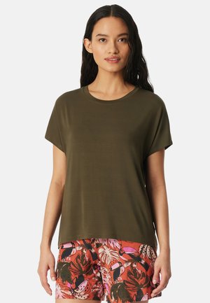 Vrouw met lang donker haar, gekleed in een olijfgroen T-shirt met korte mouwen en kleurrijke korte broek met tropische print, tegen een effen achtergrond.