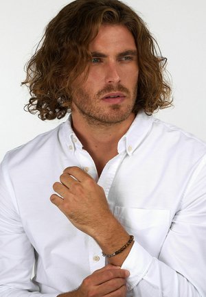 Camicia bianca con bottoni e taschino, indossata da un uomo che mostra un braccialetto con perle marroni e nere, su uno sfondo neutro.