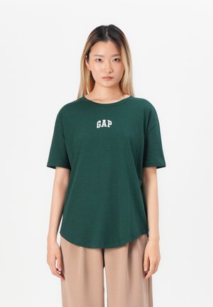 Jeune femme avec de longs cheveux portant un t-shirt GAP vert foncé et un pantalon large beige, se tenant contre un fond blanc.