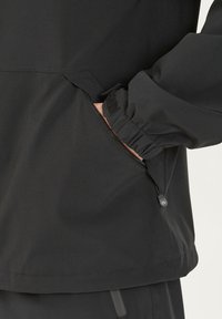 Sort jakke med en lynlåslomme, justerbar manchet med velcrolukning og snor i kanten for en sikker pasform. Glat, letvægtigt stof.