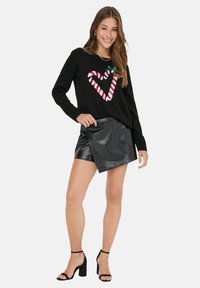 Schwarzer Pullover mit einem gestreiften Herzdesign in Rot und Weiß, kombiniert mit hochgeschnittenen schwarzen Shorts aus Kunstleder und schwarzen Sandalen mit Blockabsatz.