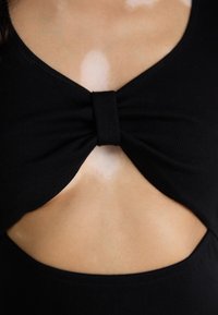 Svart ribbestrikket topp med utskjæringsdesign og en sentral sløyfedetalj, som fremhever hudtonen under stoffet.