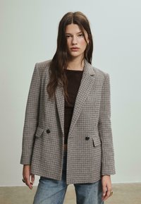 Veste à carreaux marron et beige, dotée d'un design à double boutonnage, de revers crantés et de deux poches avant. Tissu texturé.