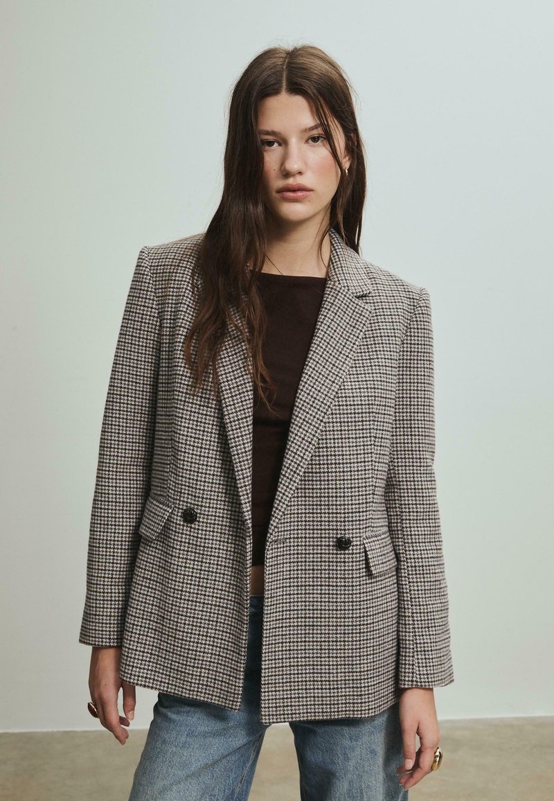 Veste à carreaux marron et beige, dotée d'un design à double boutonnage, de revers crantés et de deux poches avant. Tissu texturé.