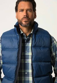 Tummansininen puhvivesti, jossa on korkea kaulus, vetoketju ja tikattu pinta, päällä sinivihreäruudullinen paita.