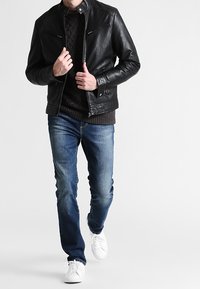 Chaqueta de cuero negra con bolsillos con cremallera en el pecho, llevada sobre un suéter de punto oscuro, combinada con jeans azules y zapatillas blancas.