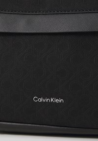 Fekete textúrájú anyag, amely dombornyomott mintával rendelkezik, és az alján egy fémes "Calvin Klein" logó található. Felső része sima bőr részletekkel van díszítve.