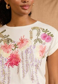Fehér, rövid ujjú felső, bonyolult virágos hímzéssel rózsaszín, piros és lila színekben, zöld levelekkel és puha textúrával.