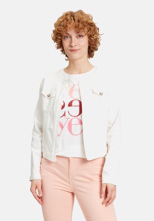 Jonge vrouw met krullend rood haar, gekleed in een witte cropped jack, een wit graphic t-shirt en lichtroze broek, staand tegen een effen achtergrond.