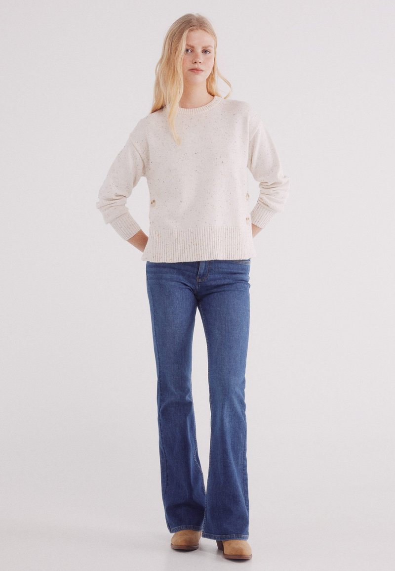 Lichtbeige gespikkelde trui met een geribbelde zoom en manchetten, gecombineerd met donkerblauwe flared jeans en tan gekleurde heeled boots. Eenvoudige, casual look.