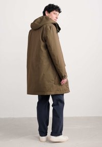 Vyras su garbanotais tamsiais plaukais, dėvintis rudą gobtuvą turinčią paltą, tamsias kelnes ir baltus sportbačius, stovi prieš paprastą šviesią фонą.