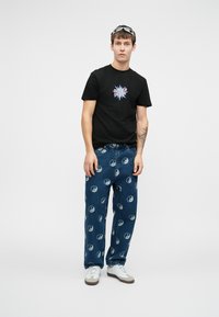 Noor mees kannab musta Santa Cruz t-särki, siniseid teksaseid valgete yin-yangi sümbolitega, valgeid tennisetossusid ja prille peas, seistes ühetaolise tausta ees.