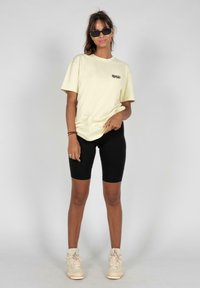 Camiseta de algodón amarillo claro con texto negro, combinada con pantalones cortos de ciclismo negros y zapatillas beige, llevada por una persona en un fondo neutro.