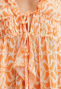 Tissu à motifs orange avec nouage à l'avant et taille froncée. Matière légère, semi-transparente, avec un motif floral et des bords volantés.