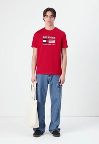 Červené bavlněné tričko s logem "HILFIGER" a datem založení. V kombinaci se světle modrými volnými džíny a černými botami, drží krémovou tašku.