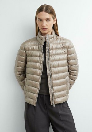 Frau trägt eine hellbeige gesteppte Daunenjacke über einem dunklen Oberteil und lockeren dunklen Hosen, stehend vor einem einfarbigen weißen Hintergrund.