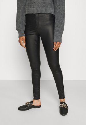 Persoon draagt zwarte legging van nep leer, grijze gebreide trui en zwarte instap loafers met een grote gouden ketting, staand op een grijze vloer.