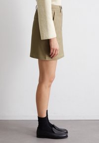 Jupe en coton khaki avec coupe droite et poches, associée à des mocassins noirs et des chaussettes foncées. Design simple, sans motifs ni accents.