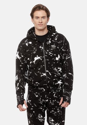 Młody mężczyzna ubrany w czarną bluzę na zamek i spodnie w białe plamy farby, stojący na tle jednolitego białego tła.