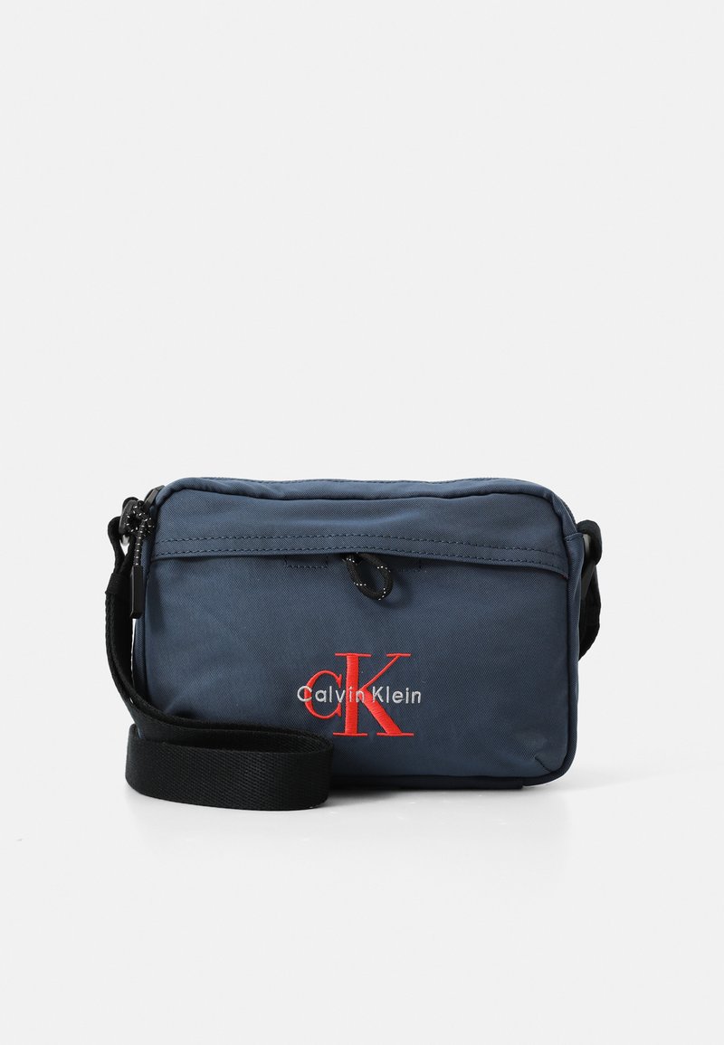 Bolso cruzado de nylon azul marino con cierre de cremallera, que presenta un logo bordado en rojo, correa ajustable y detalle en el bolsillo frontal.