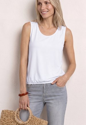 Femme souriante portant un haut blanc sans manches et un pantalon bleu rayé tenant un sac à main tissé avec des poignées rondes devant un mur clair.