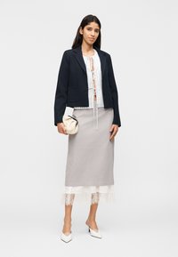 Γυναίκα που φορά μαύρο σακάκι πάνω από λευκό μπλουζάκι με δετό μπροστινό μέρος, γκρι φούστα με δαντελωτό τελείωμα, λευκά ψηλοτάκουνα παπούτσια, κρατώντας μικρό μπεζ τσαντάκι, στέκεται μπροστά σε μονόχρωμο φόντο.