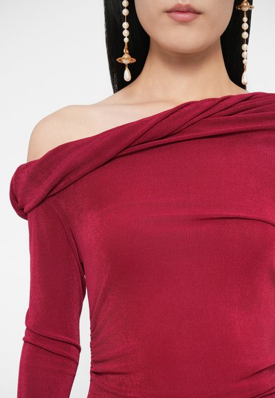 Sieviete tērpusies tumši sarkanā off-plecu topā ar garām piedurknēm un nēsā garas zelta auskarus, rotātus ar pērlēm, uz vienkārša fona.