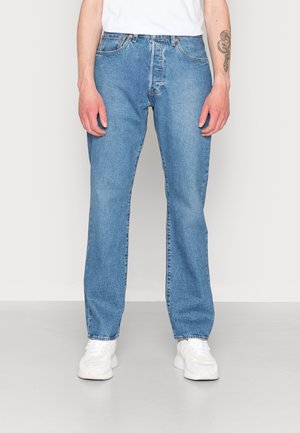 Personne portant un jean droit bleu clair, des baskets blanches et une chemise blanche, debout devant un fond clair uni.