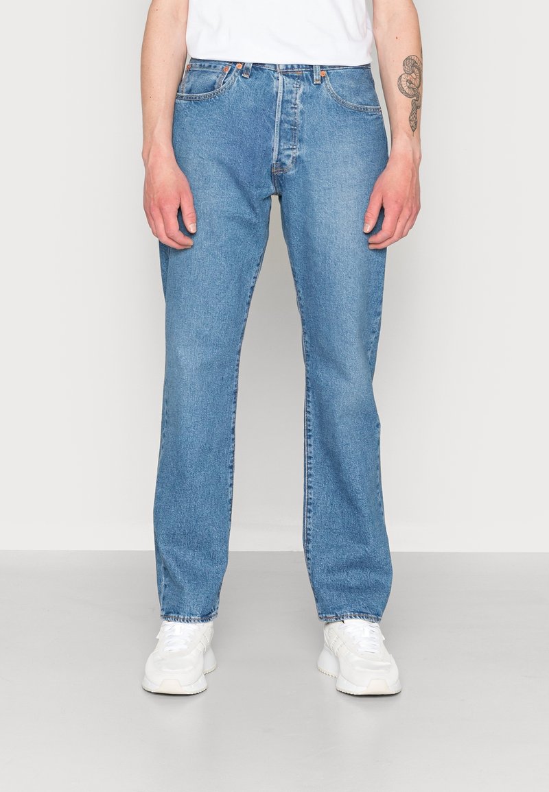 Personne portant un jean droit bleu clair, des baskets blanches et une chemise blanche, debout devant un fond clair uni.