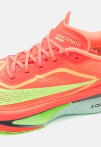 Nike Zoom Fly juoksukengät kirkkaan korallinvärisinä vihreillä yksityiskohdilla; ominaisuuksia ovat verkko-osa, kerroksellinen muotoilu ja voimakkaasti kaareva välipohja.