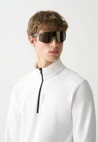 Bela flis majica s zip-kragnico in črnimi detajli, v kombinaciji z oversized temnimi ogledali očali z geometrijskim vzorcem.