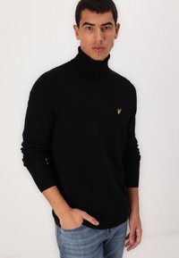 Schwarzer Rollkragenpullover aus weichem, strukturiertem Stoff mit einem figurnahen Design. Mit einem kleinen gelben Logo auf der linken Brust.
