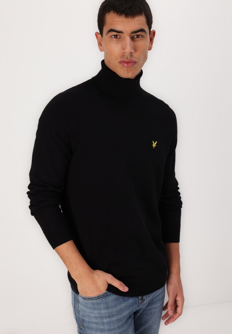Pull à col roulé noir en tissu doux et texturé avec une coupe ajustée. Présente un petit logo jaune sur la poitrine gauche.